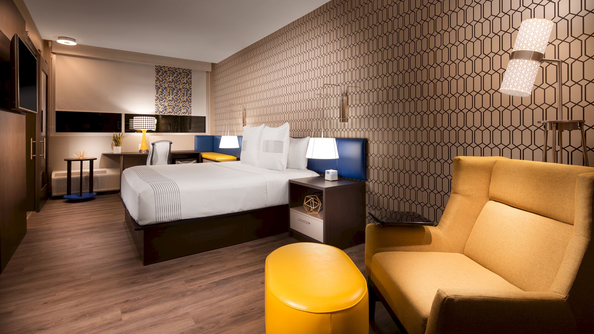 A modern hotel room with a bed, yellow armchair, desk, and geometric-patterned wallpaper. The room features warm lighting and contemporary decor.