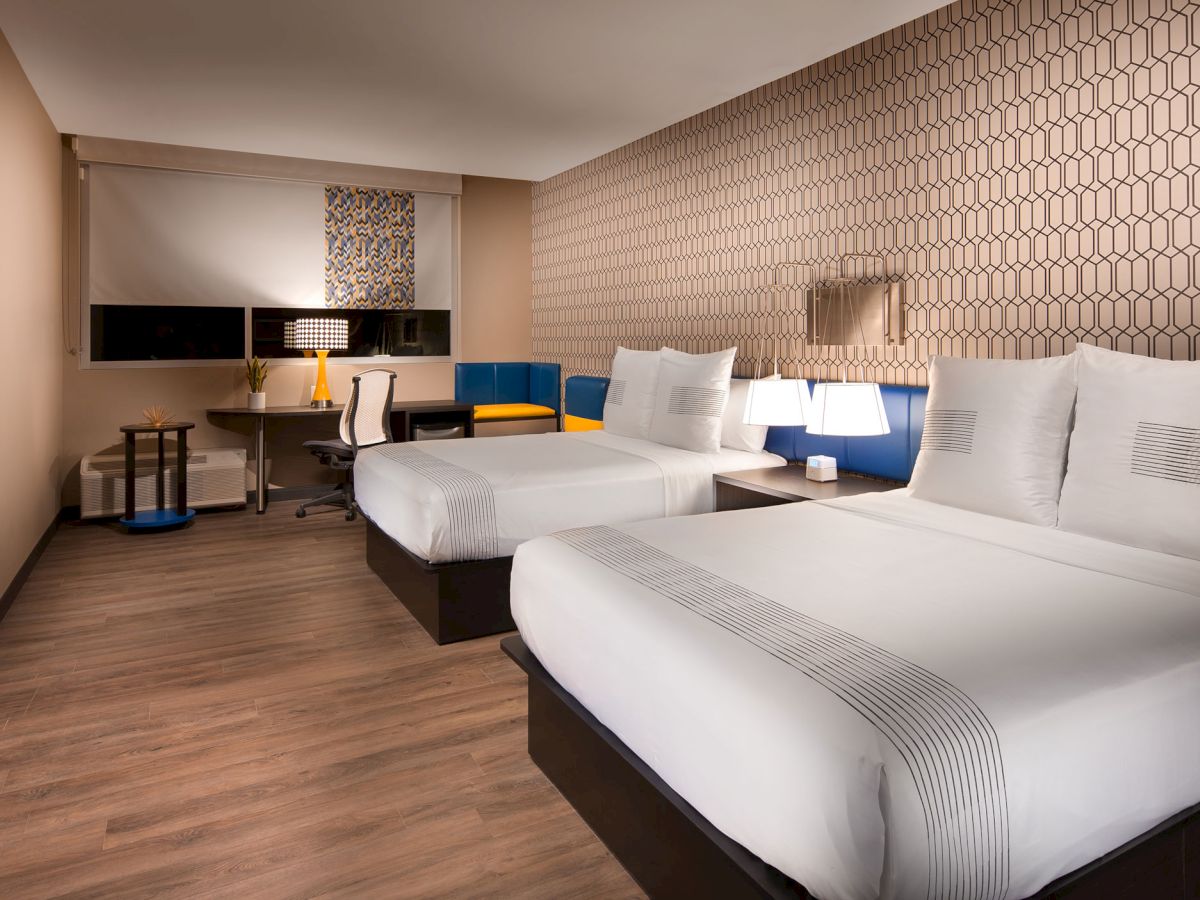 A modern hotel room with two beds, a wall-mounted TV, a desk with a chair and lamp, and geometric wallpaper.