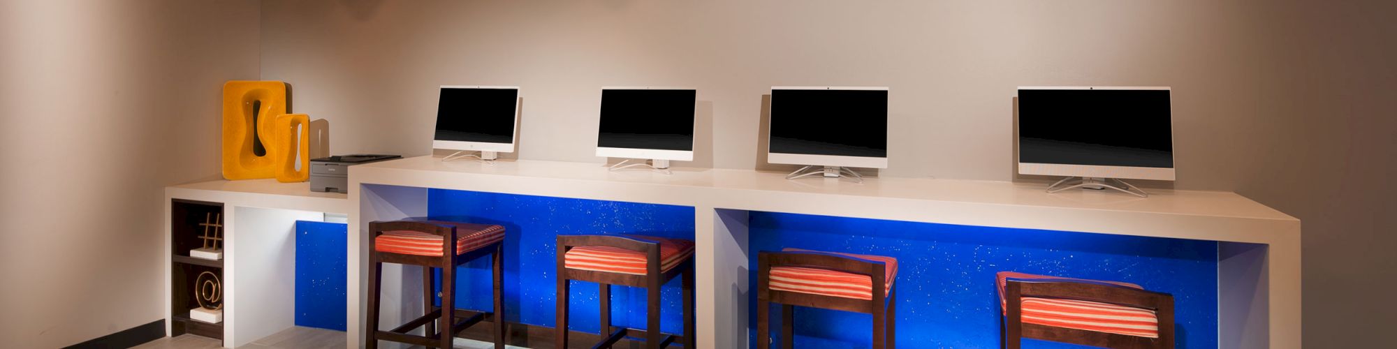 The image shows a modern computer lab with four monitors, three stools, and hanging light fixtures, with a blue backdrop and minimal decor ending the sentence.