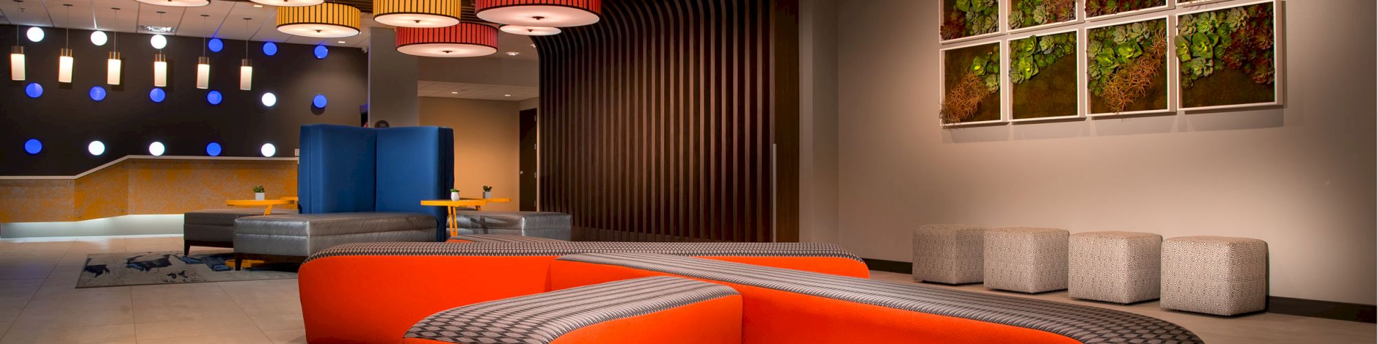 The image shows a modern lobby with vibrant orange seating, colorful ceiling lights, wall art, and a contemporary decor style, with seating cubes aligned.