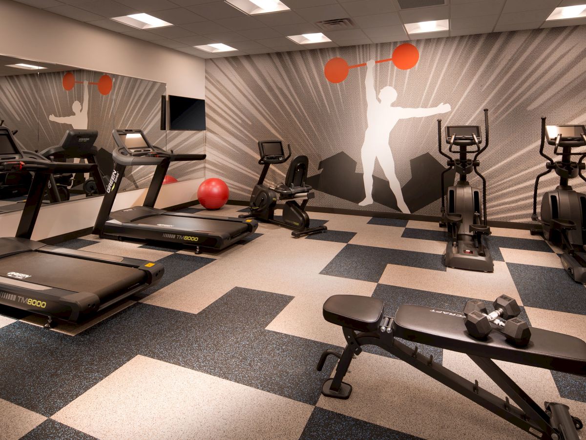 The image shows a gym with treadmills, exercise bikes, a bench with dumbbells, a red exercise ball, and a wall mural of a weightlifter, ending the sentence.