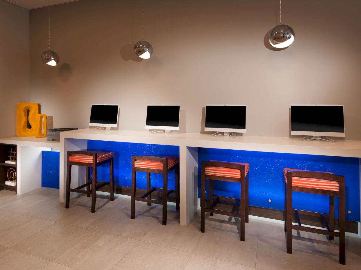 A modern workspace with four computers on a white counter, each paired with a stool. Overhead lighting and orange accents create a cozy atmosphere.