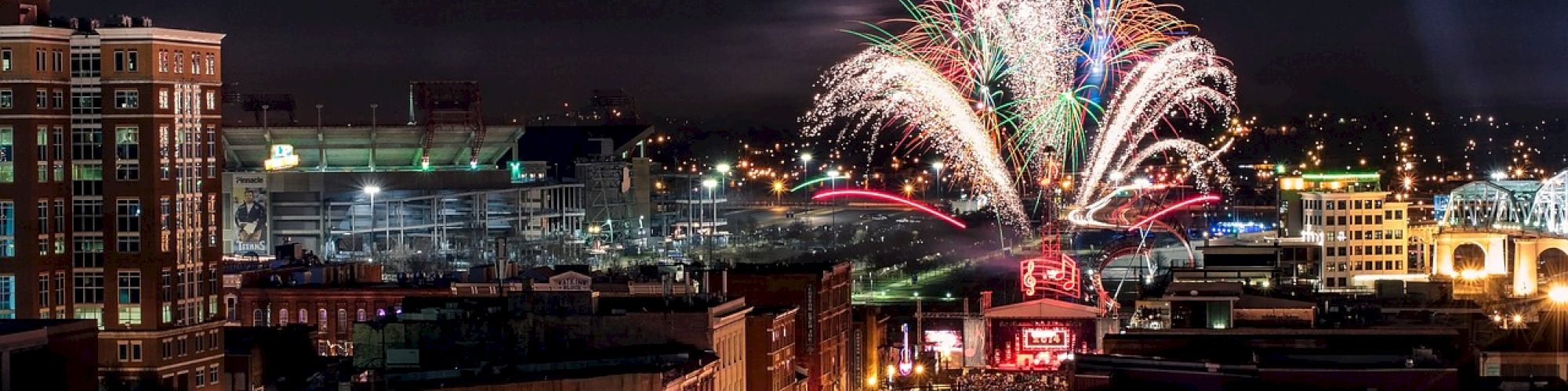 A bustling city street at night with fireworks lighting up the sky, surrounded by buildings, and a large crowd of people below.