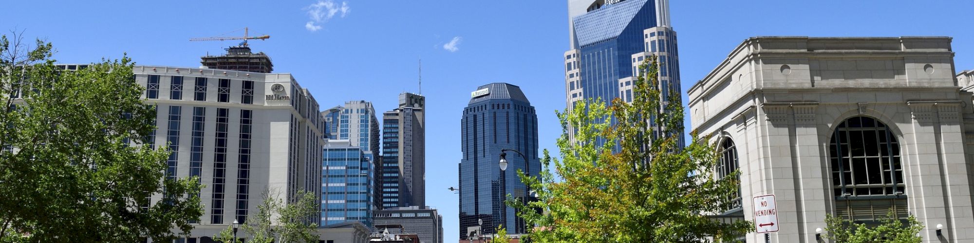 A city street with vehicles, including a bus, and pedestrians, under a blue sky. The street sign reads 