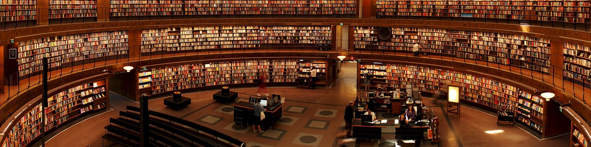 The image shows a large, circular, multi-level library with rows of bookshelves and reading tables in the center. There are people reading.