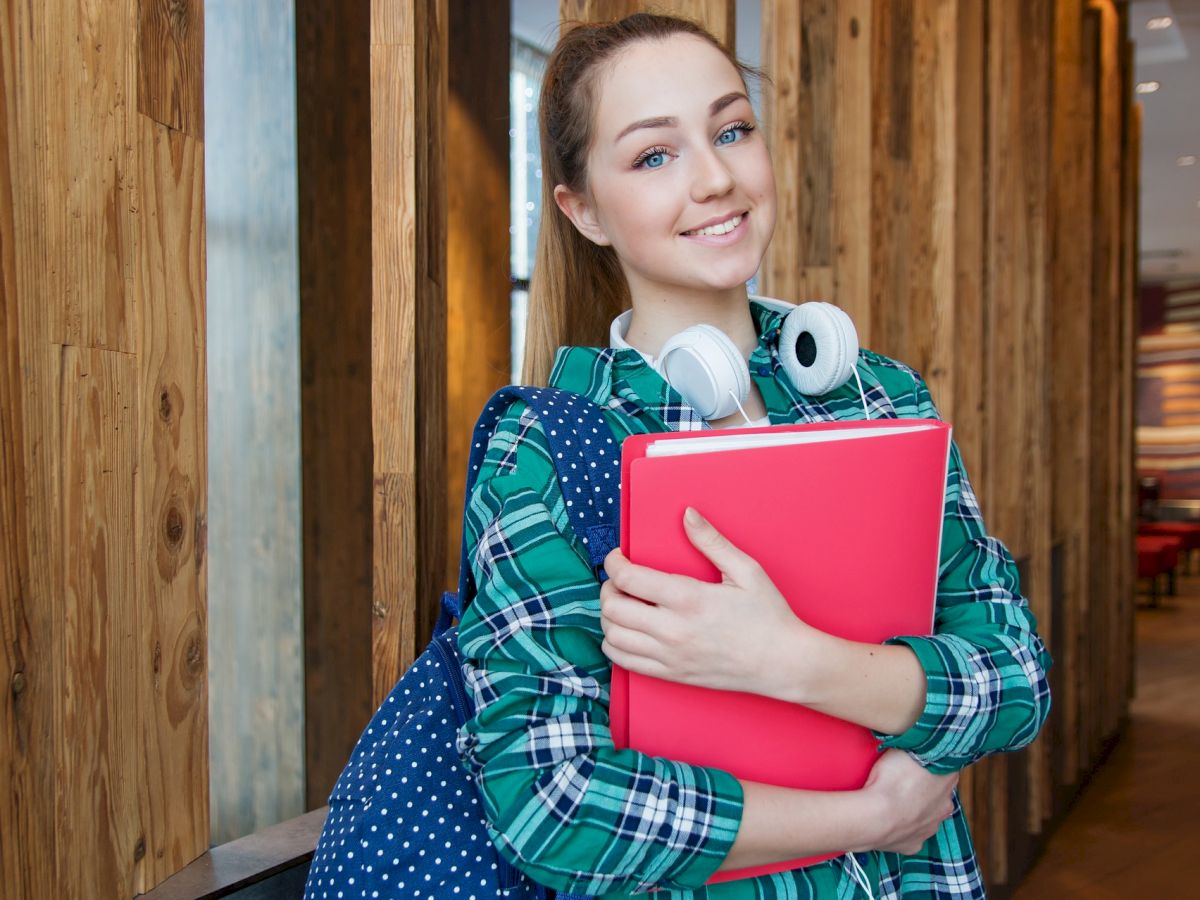 A person with red hair smiles, holding a pink folder, wearing headphones around the neck, and carrying a blue polka-dot bag in a hallway.