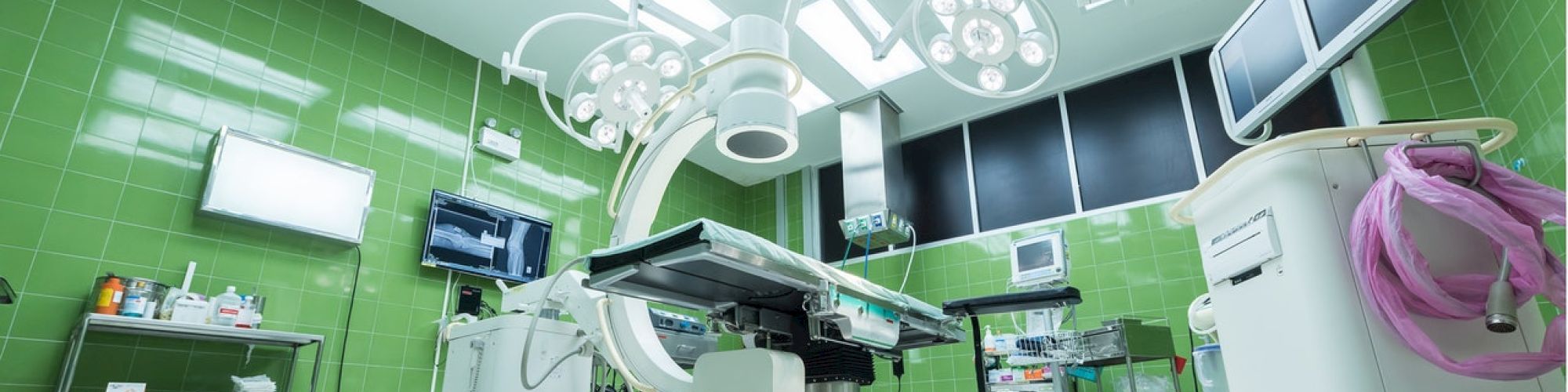 The image shows a modern, well-equipped operating room with green tiled walls, an operating table, surgical lights, and various medical equipment.