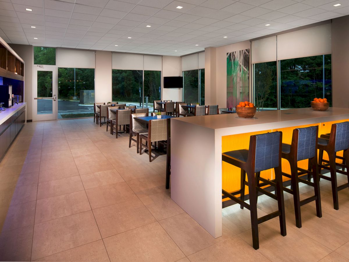 A modern dining area with high chairs around a glowing counter, multiple tables and chairs, large windows, and a serving counter on the left.