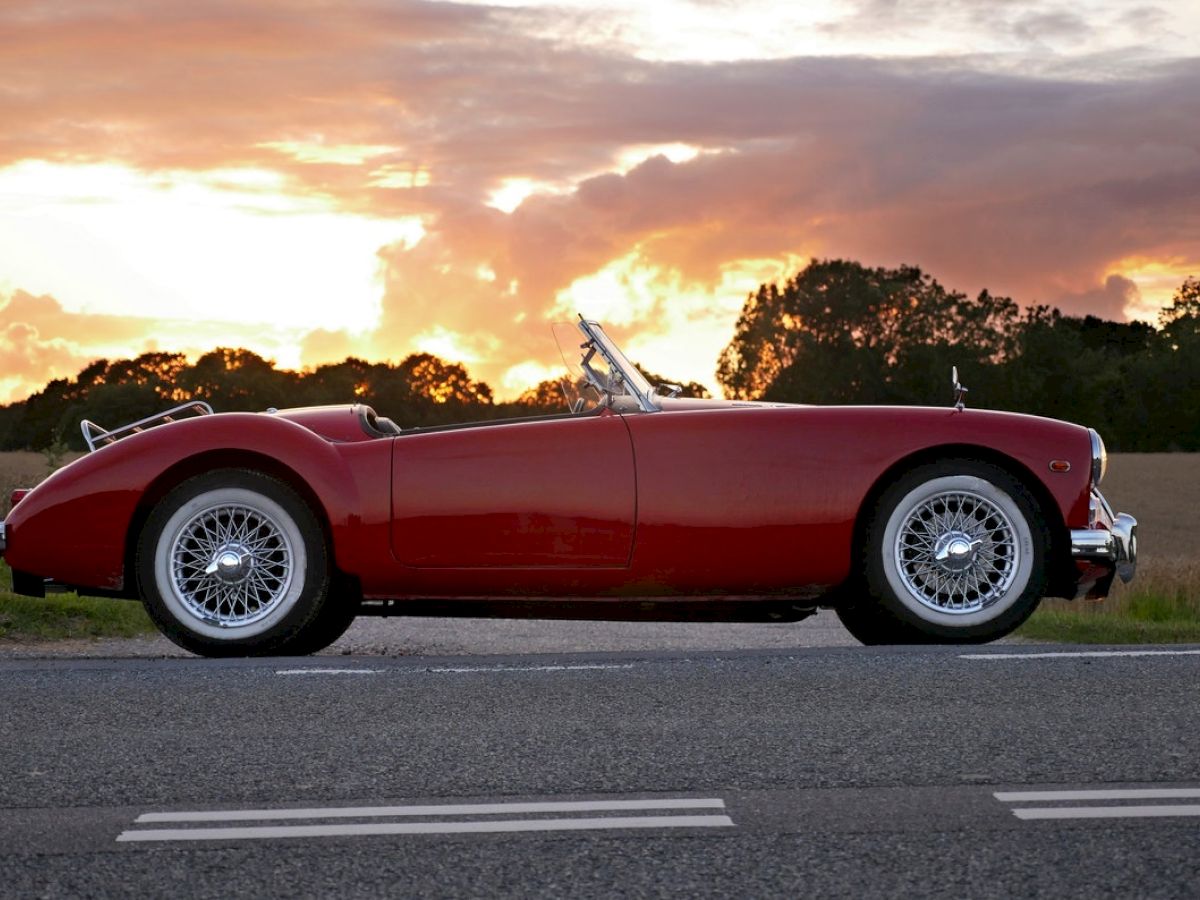 A red vintage convertible car is parked by the roadside with a beautiful sunset and countryside backdrop in the background.