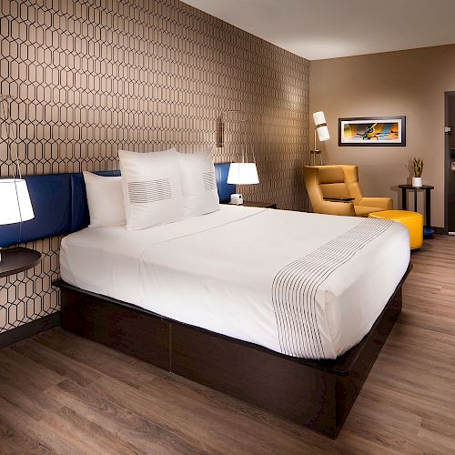 This image shows a modern hotel room with a large bed, nightstands, lamps, a yellow armchair, a wall-mounted TV, and a patterned wall accent.