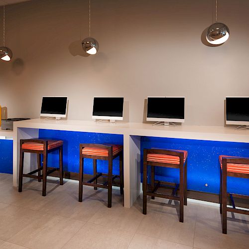 The image shows a modern computer lab with four computers on a white desk, wooden stools with red cushions, and ambient lighting.