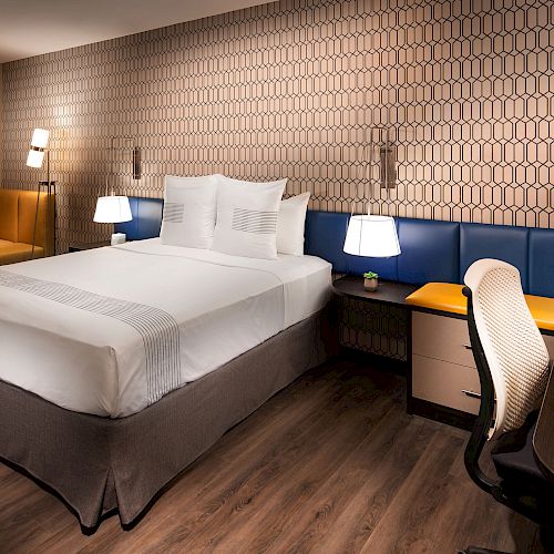 A modern hotel room with a double bed, desk and chair, corner seating, stylish lamps, patterned wallpaper, and wood flooring.