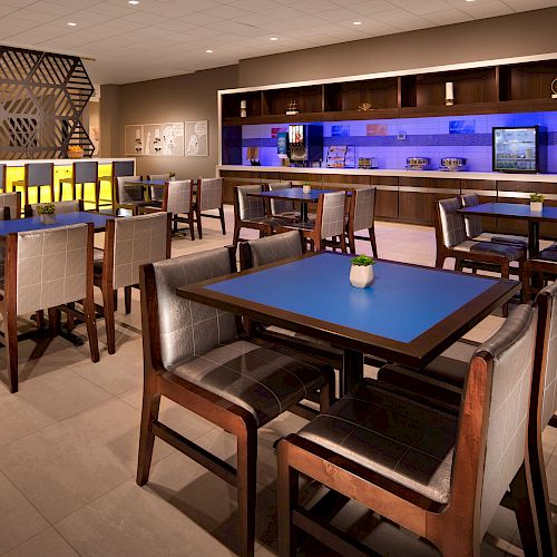 The image shows a modern dining area with tables, chairs, a counter with food and drink amenities, and blue accent lighting on the back wall.