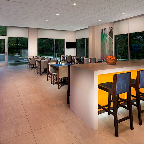 This image shows a modern kitchen area with a long island, bar stools, cabinets, a countertop, dining tables, and large windows with a view outside.