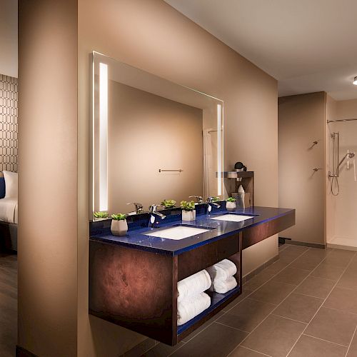 The image shows a modern hotel room with a bathroom vanity, a shower area, and part of a bedroom with a bedside table and lamp.