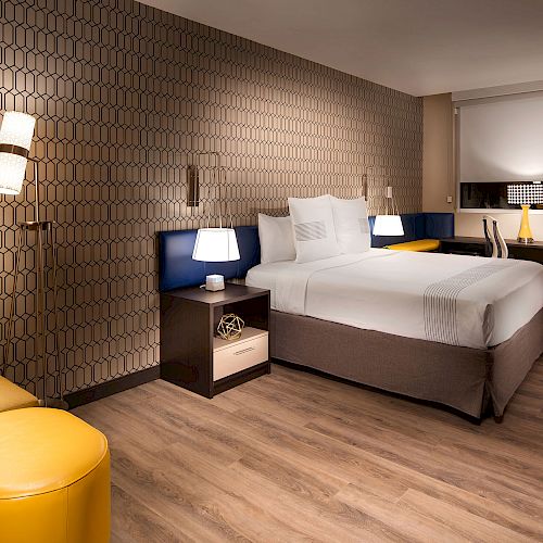 This image shows a modern hotel room with a king-size bed, nightstands, lamps, a sofa, a small table, and stylish decor on the walls and floor.