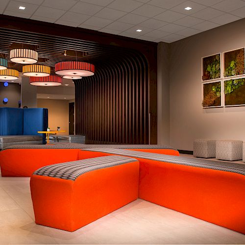 A modern lobby with orange seats, colorful hanging lights, wall art, and stools by the wall. The room features a bright and contemporary design ending the sentence.