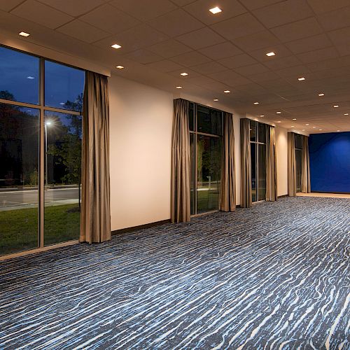 An empty, well-lit room has large windows with curtains, a patterned carpet, and a blue accent wall with a mounted flat-screen TV.