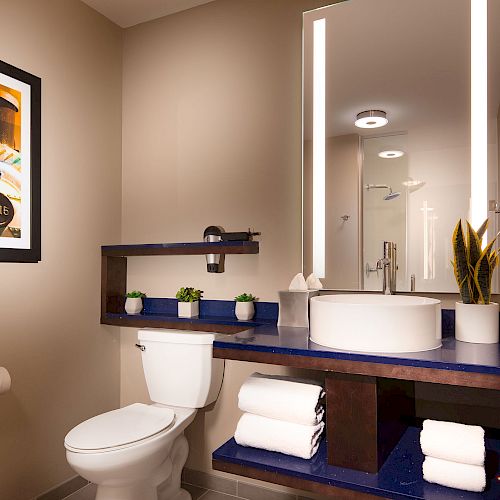 A modern bathroom with a toilet, sink, large mirror, wall art, and shelves holding towels and plants.