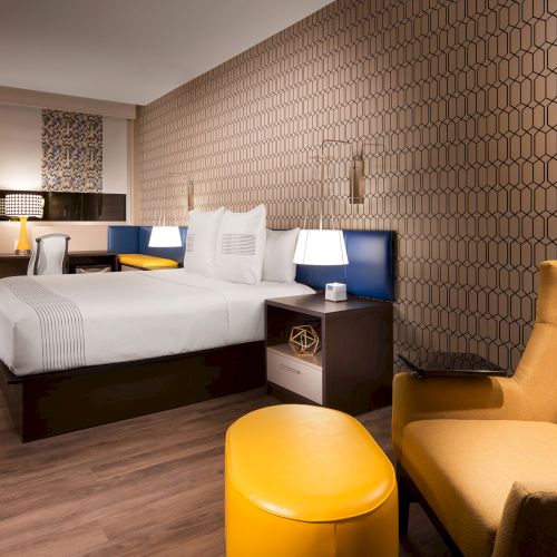 A modern hotel room with a double bed, yellow seating, a desk, and ambient lighting. The decor features a hexagonal patterned wall.