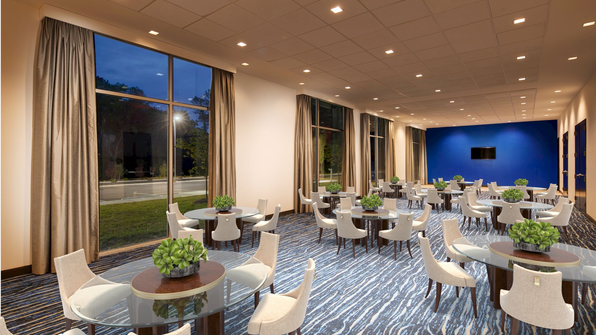 The image shows a modern dining area with round tables, white chairs, green centerpieces, large windows with curtains, blue carpeting, and a TV on a blue wall.